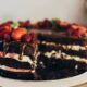 Bolo de chocolate com camadas, recheio branco/rosa, calda, morangos, framboesas, uma fatia removida