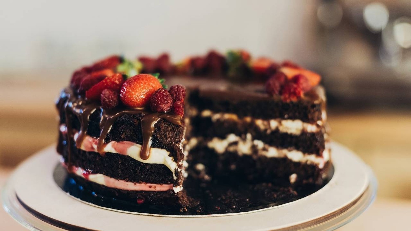 Bolo de chocolate com camadas, recheio branco/rosa, calda, morangos, framboesas, uma fatia removida