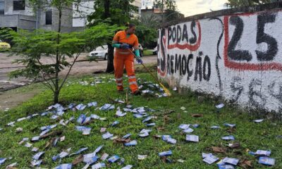 Uma gari de uniforme laranja varre panfletos eleitorais do gramado ao lado de um muro pichado