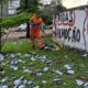Uma gari de uniforme laranja varre panfletos eleitorais do gramado ao lado de um muro pichado