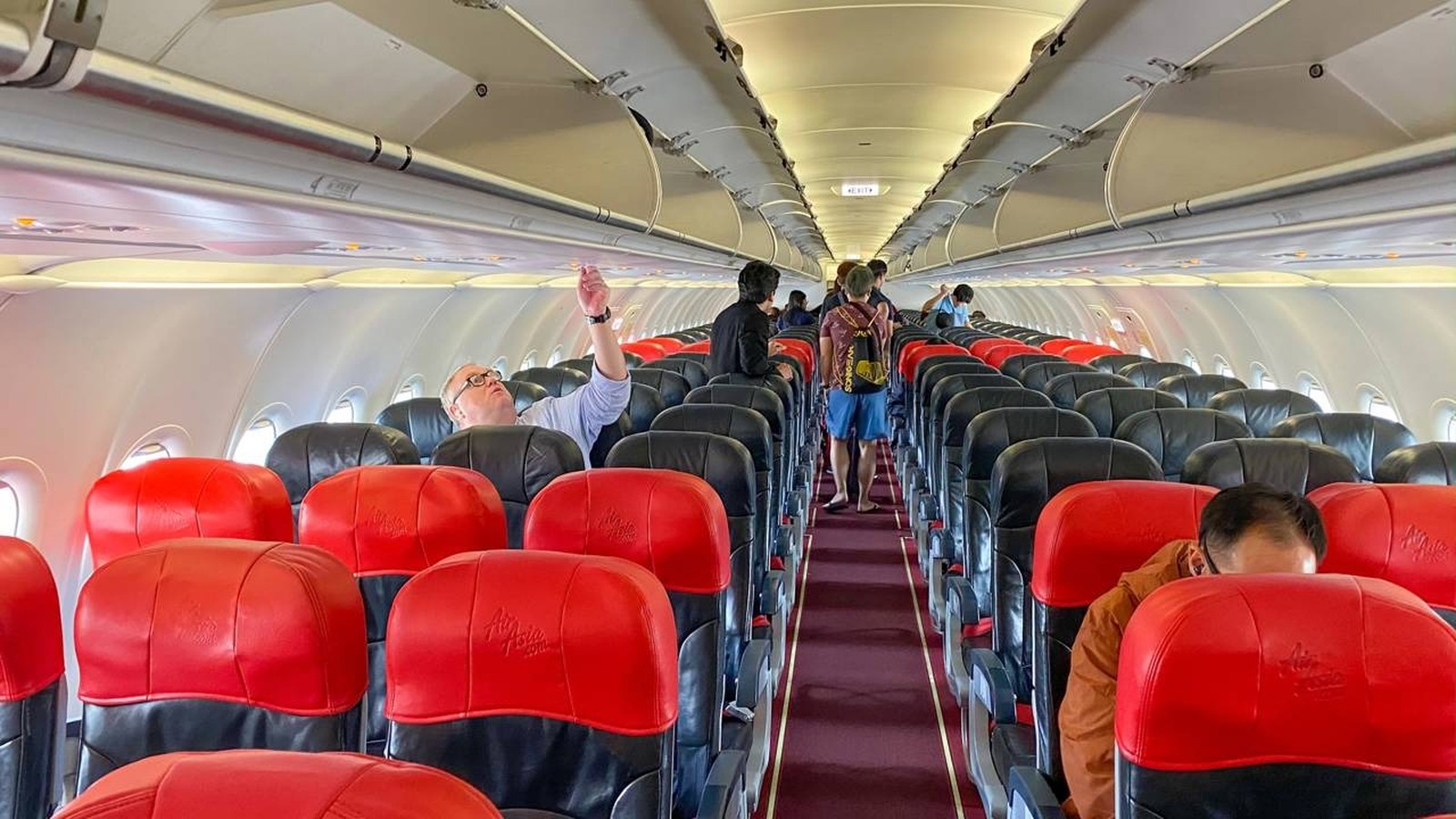 Interior de aeronave AirAsia com assentos vermelhos e pretos, passageiros e compartimentos de bagagem abertos