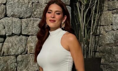 Ana Clara sorrindo, cabelo ruivo ondulado, blusa branca gola alta, brincos grandes, parede de pedras