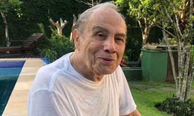 Homem idoso de cabelos grisalhos e camiseta branca sorri para câmera em área externa com piscina