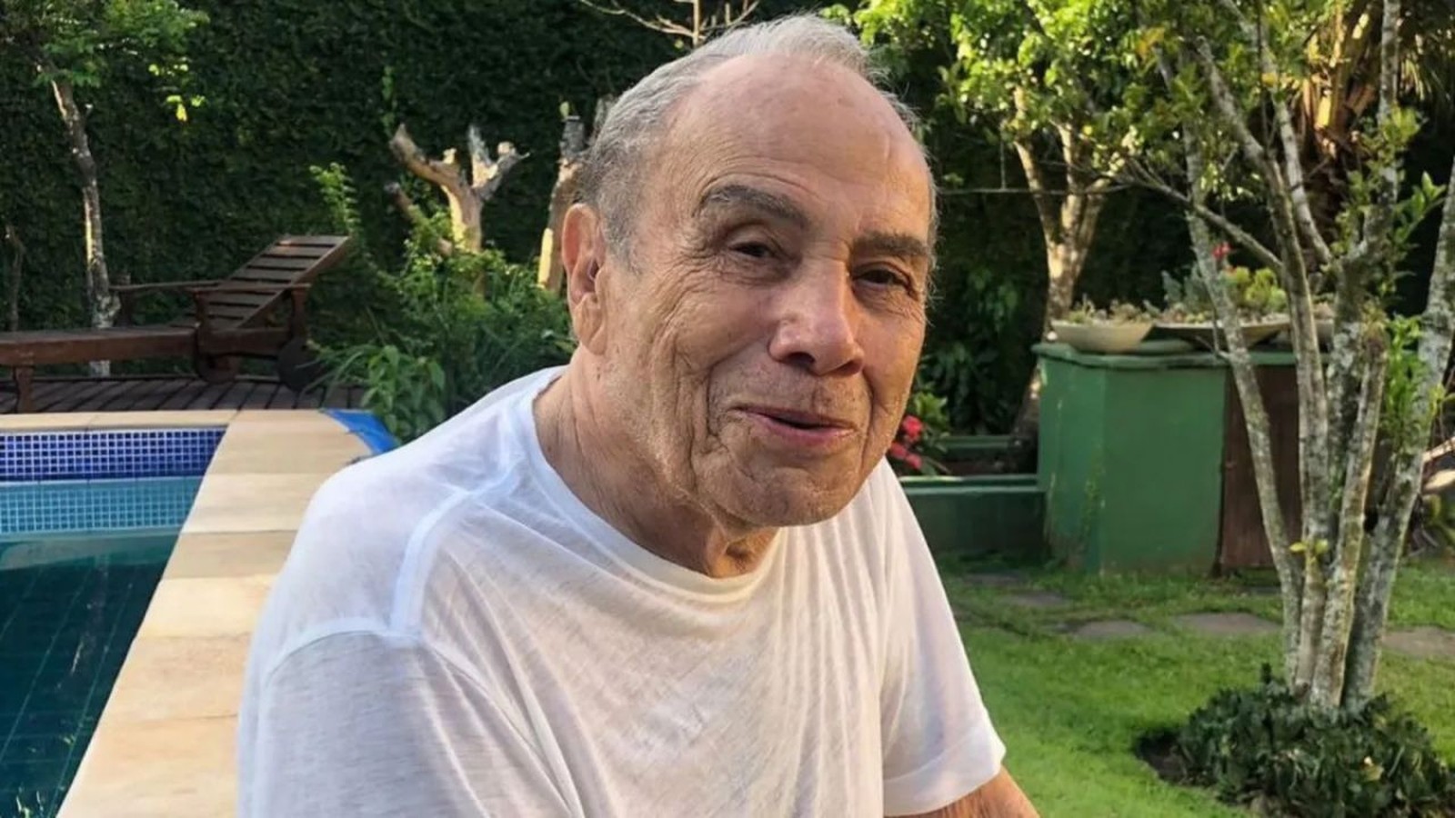 Homem idoso de cabelos grisalhos e camiseta branca sorri para câmera em área externa com piscina