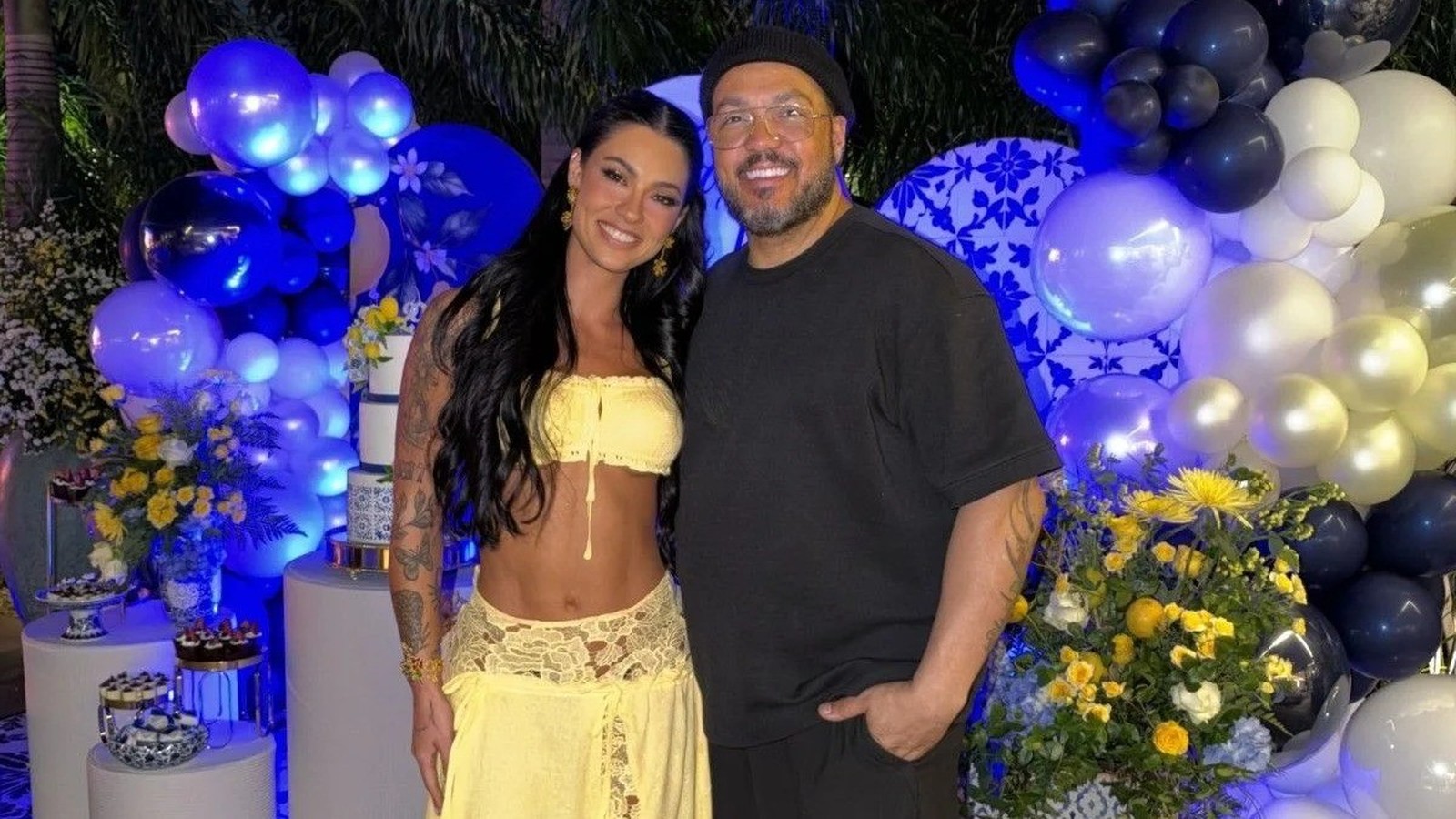 Casal sorridente Rayane Figliuzzi e Belo em festa com balões azuis e flores amarelas