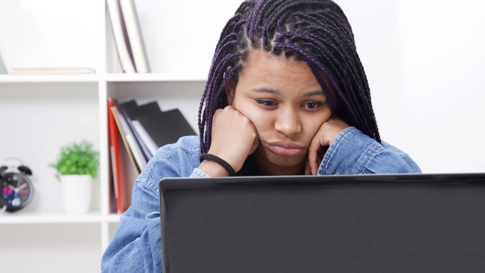 Jovem com cabelo trançado e detalhes roxos, encosta o rosto nas mãos, olhando para um laptop