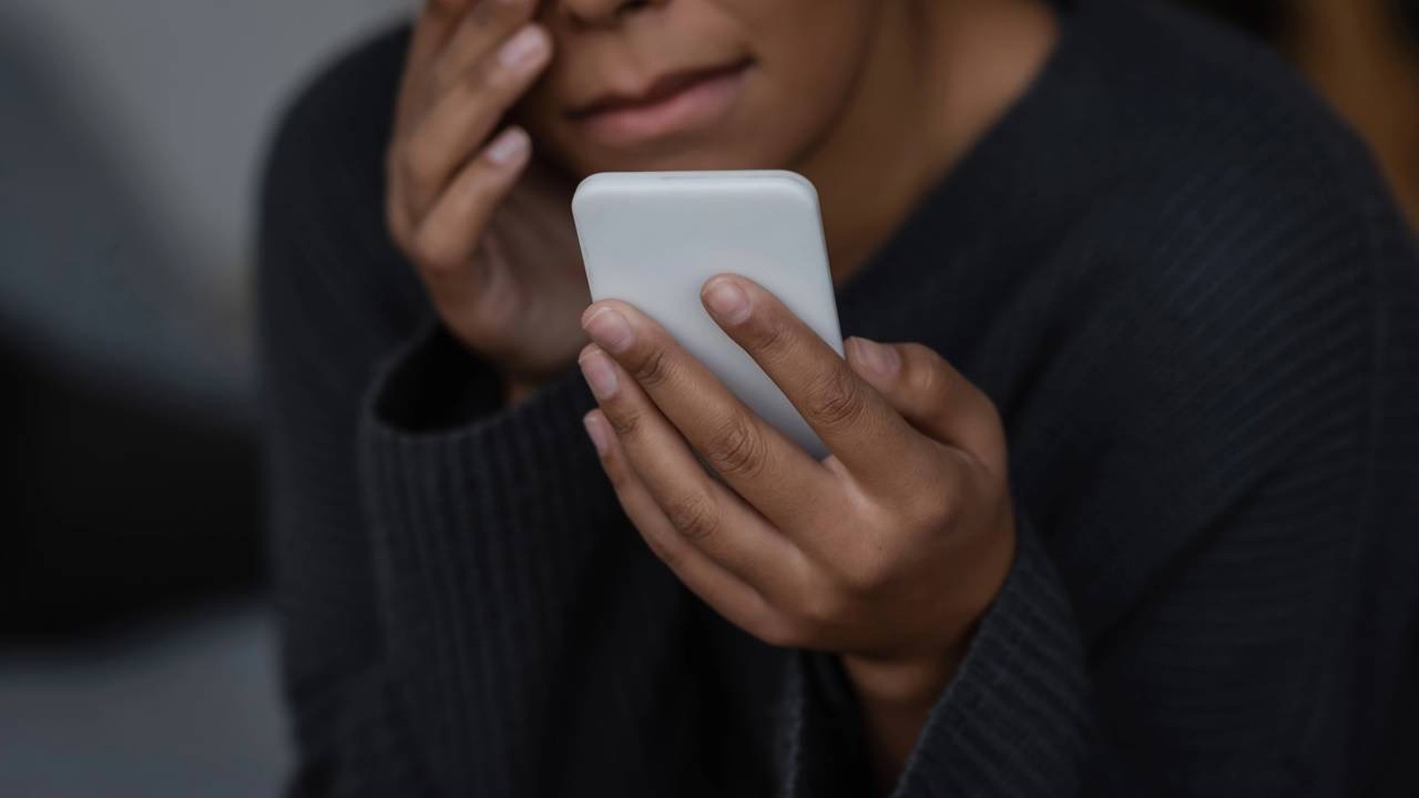 Pessoa de pele escura com suéter escuro segura um celular branco e esfrega o olho