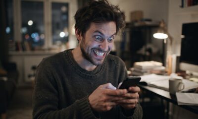 Homem barbudo com olhos arregalados e sorriso forçado olhando intensamente para um smartphone