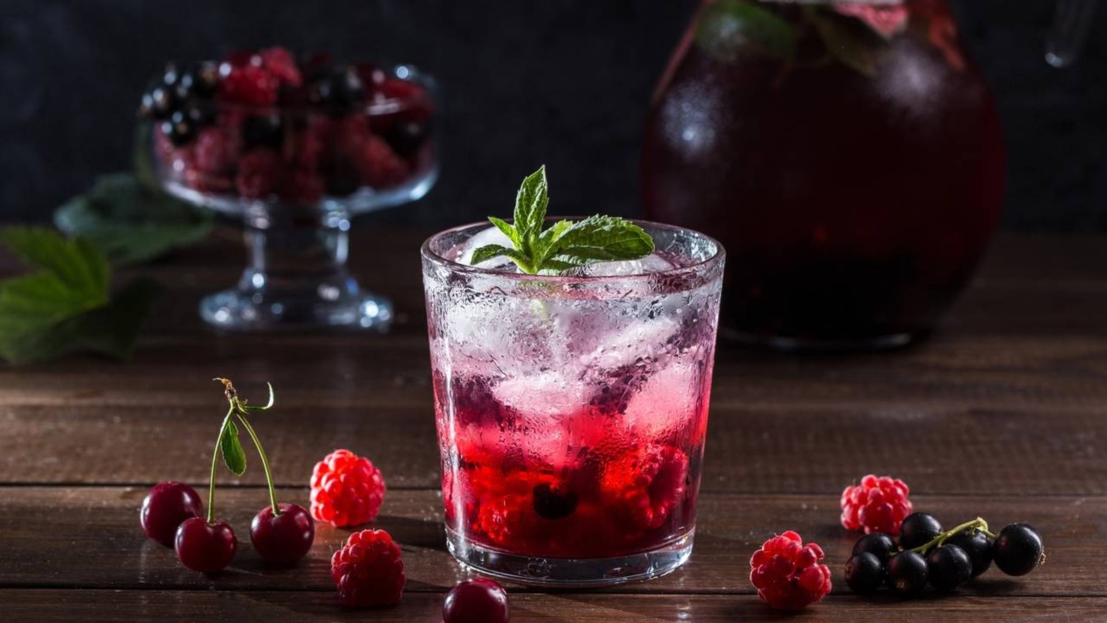 Copo com drinque de frutas vermelhas, gelo, hortelã e frutas frescas em mesa de madeira