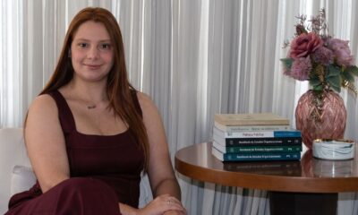 Jovem sorridente com cabelo ruivo sentada, ao lado de livros "Política Pública" e flores