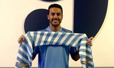 Homem sorrindo segura cachecol listrado azul e branco da Lazio, uniforme azul claro
