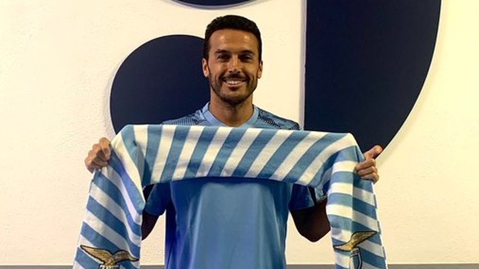 Homem sorrindo segura cachecol listrado azul e branco da Lazio, uniforme azul claro