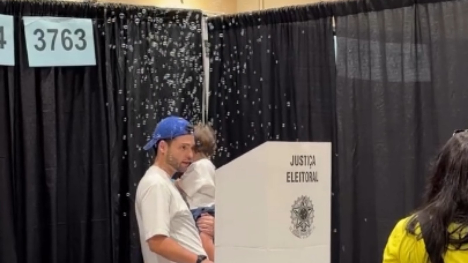 Homem com boné azul segura criança em urna eleitoral com bolhas. Texto "JUSTIÇA ELEITORAL" e "3763".