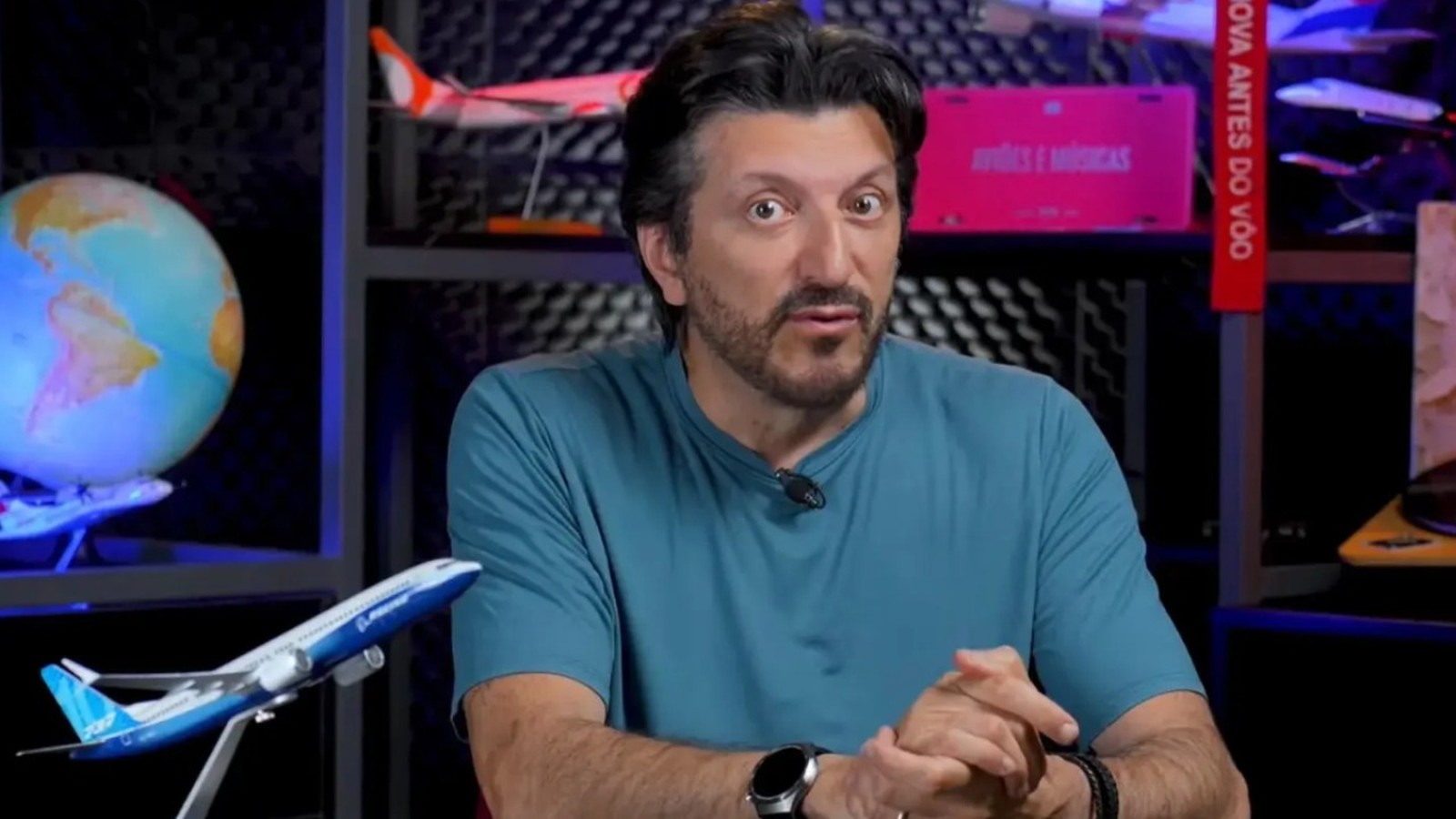 Homem de barba em camiseta teal, em estúdio com modelos de avião, globo e "Aviões e Músicas"