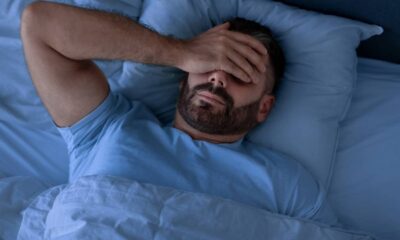 Homem barbudo, de camiseta azul, deitado na cama, com a mão sobre os olhos