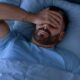 Homem barbudo, de camiseta azul, deitado na cama, com a mão sobre os olhos