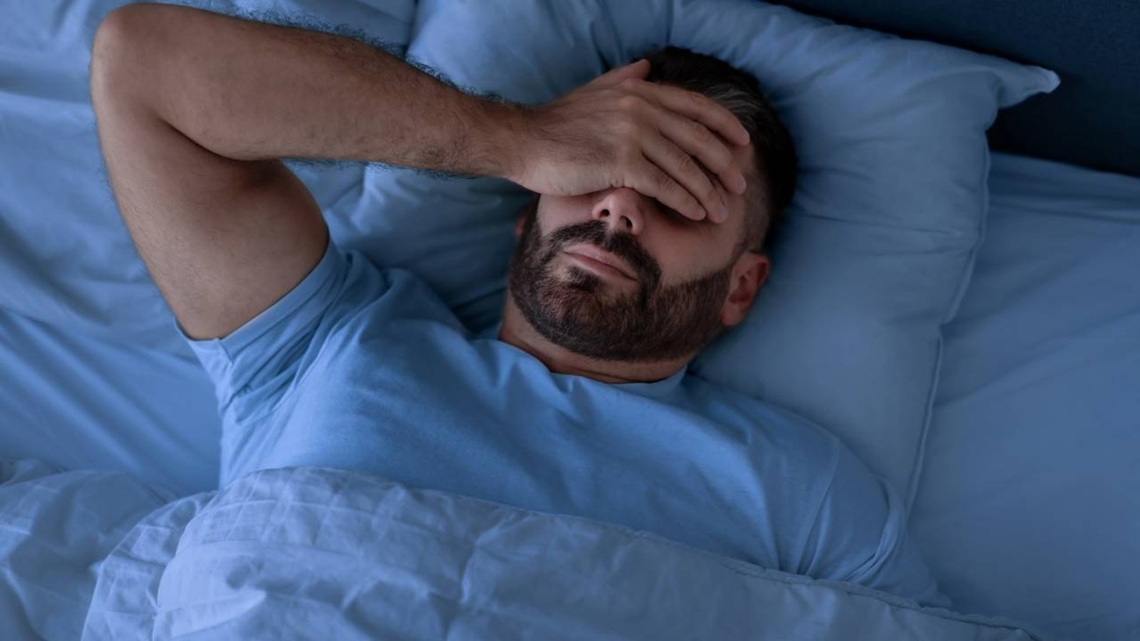 Homem barbudo, de camiseta azul, deitado na cama, com a mão sobre os olhos