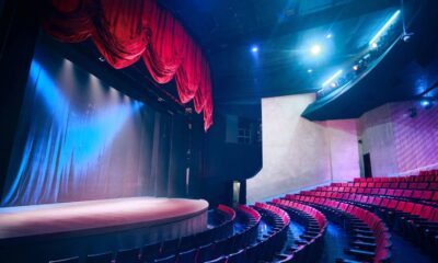 Interior de um teatro vazio com palco de madeira iluminado e cortinas vermelhas, e poltronas vermelhas.