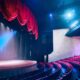 Interior de um teatro vazio com palco de madeira iluminado e cortinas vermelhas, e poltronas vermelhas.