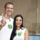 Douglas Ruas e Mariana Barbosa sorrindo, ele faz "V", com caixa "JUSTIC ELEITOF" no fundo.