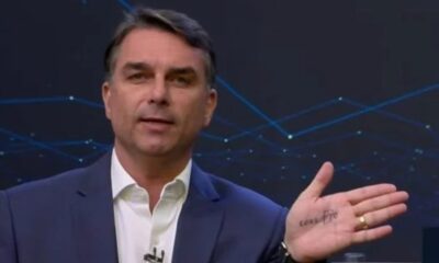 Flávio Bolsonaro, em terno azul, gesticula mostrando a palma da mão esquerda com "corrupto" escrito.