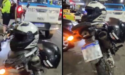 Parte traseira de uma motocicleta com dois capacetes e placa brasileira, com viatura policial ao fundo