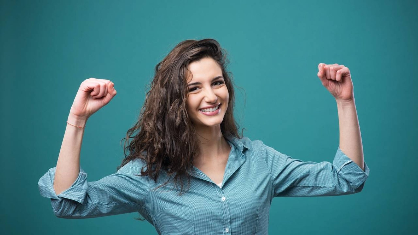 Mulher sorridente de cabelo escuro, em camisa azul, flexiona os braços com punhos cerrados