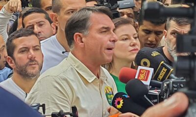Flávio Bolsonaro, camisa clara, fala, microfones com "96.5", "OESTE" e pessoas ao redor