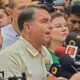 Flávio Bolsonaro, camisa clara, fala, microfones com "96.5", "OESTE" e pessoas ao redor