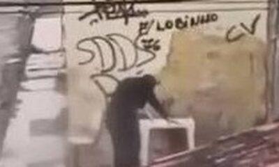 Pessoa abaixada em frente a muro com pichações "CV" e "LOBINHO" em cena de assalto a boca de fumo