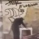 Pessoa abaixada em frente a muro com pichações "CV" e "LOBINHO" em cena de assalto a boca de fumo