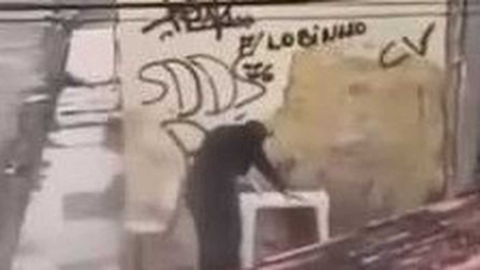 Pessoa abaixada em frente a muro com pichações "CV" e "LOBINHO" em cena de assalto a boca de fumo