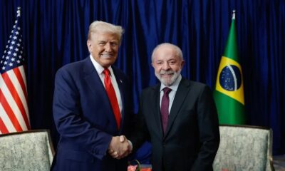 Donald Trump e Luiz Inácio Lula da Silva sorriem e apertam as mãos, com bandeiras ao fundo