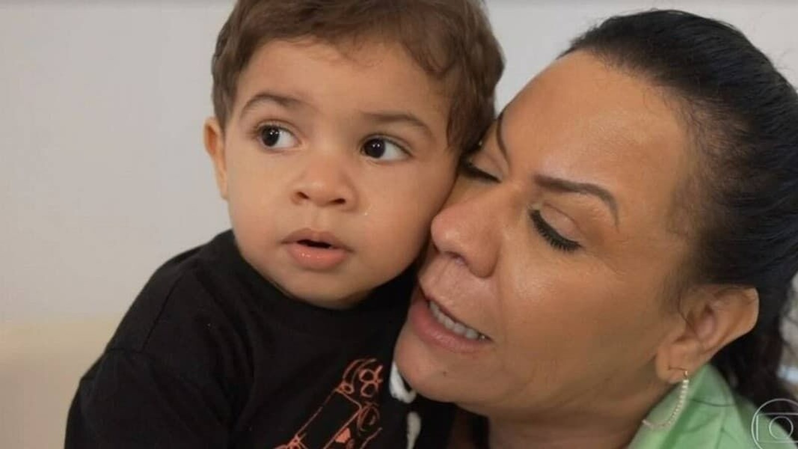 Mulher adulta com olhos fechados abraça menino pequeno com lágrima no rosto e cabelo cacheado