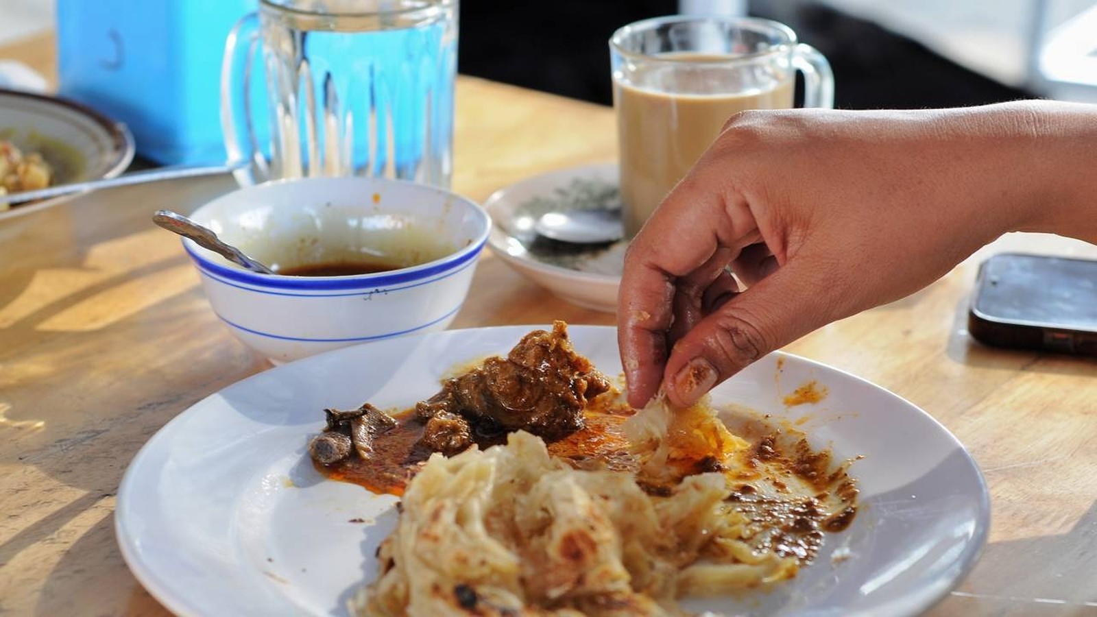 Mão pegando roti com curry em prato. Copos com água e chá, celular à mesa