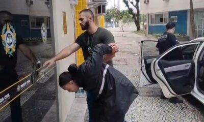 Suspeito algemado é conduzido por agente da Polícia Civil perto de carro