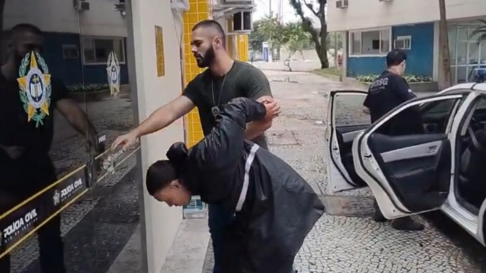 Suspeito algemado é conduzido por agente da Polícia Civil perto de carro