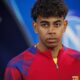 Jovem homem de cabelo encaracolado vestindo camisa vermelha e azul do Barcelona, olhando à frente