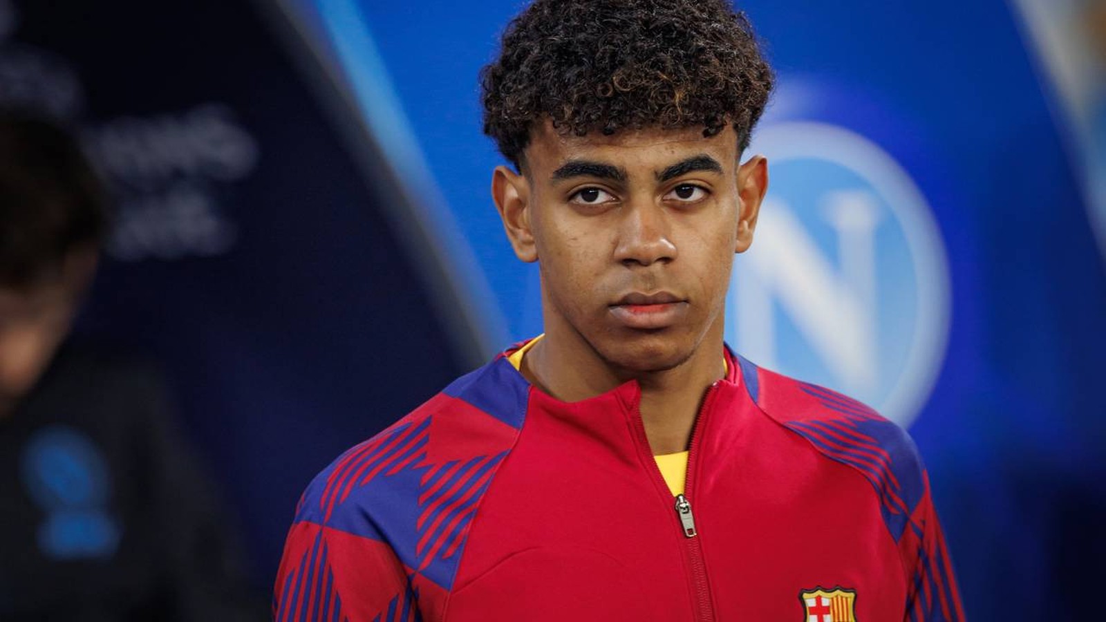 Jovem homem de cabelo encaracolado vestindo camisa vermelha e azul do Barcelona, olhando à frente