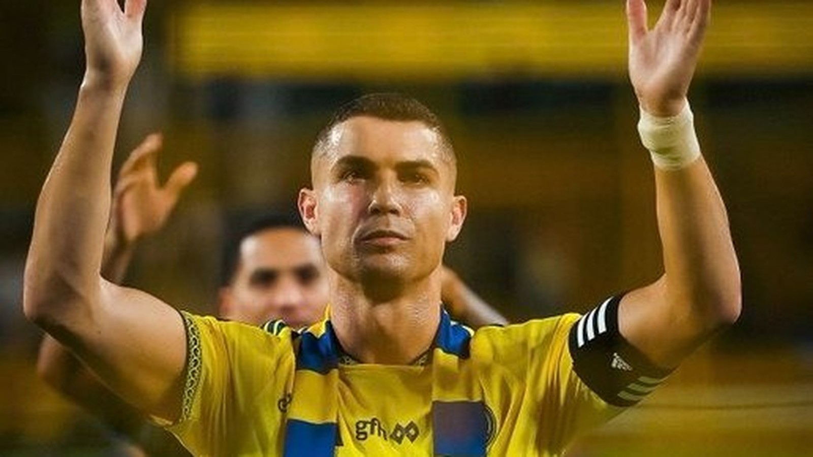 Cristiano Ronaldo com cabelo raspado, camisa amarela e azul, braços levantados com as mãos abertas