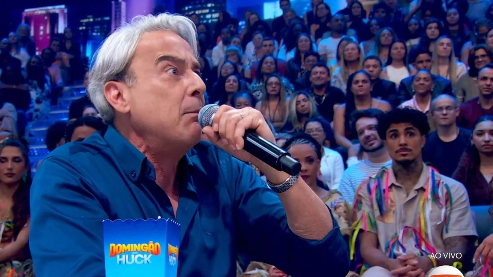 Luciano Huck, camisa azul, microfone na mão, no palco do Domingão Huck ao vivo