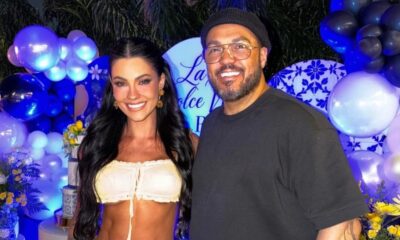 Rayane Figliuzzi e Belo, sorrindo, em festa com balões azuis e brancos, fundo com letreiro