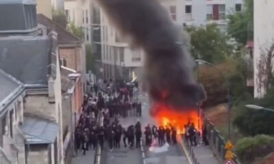 Vista aérea de uma rua com protesto estudantil, grande incêndio e fumaça escura subindo.