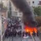 Vista aérea de uma rua com protesto estudantil, grande incêndio e fumaça escura subindo.