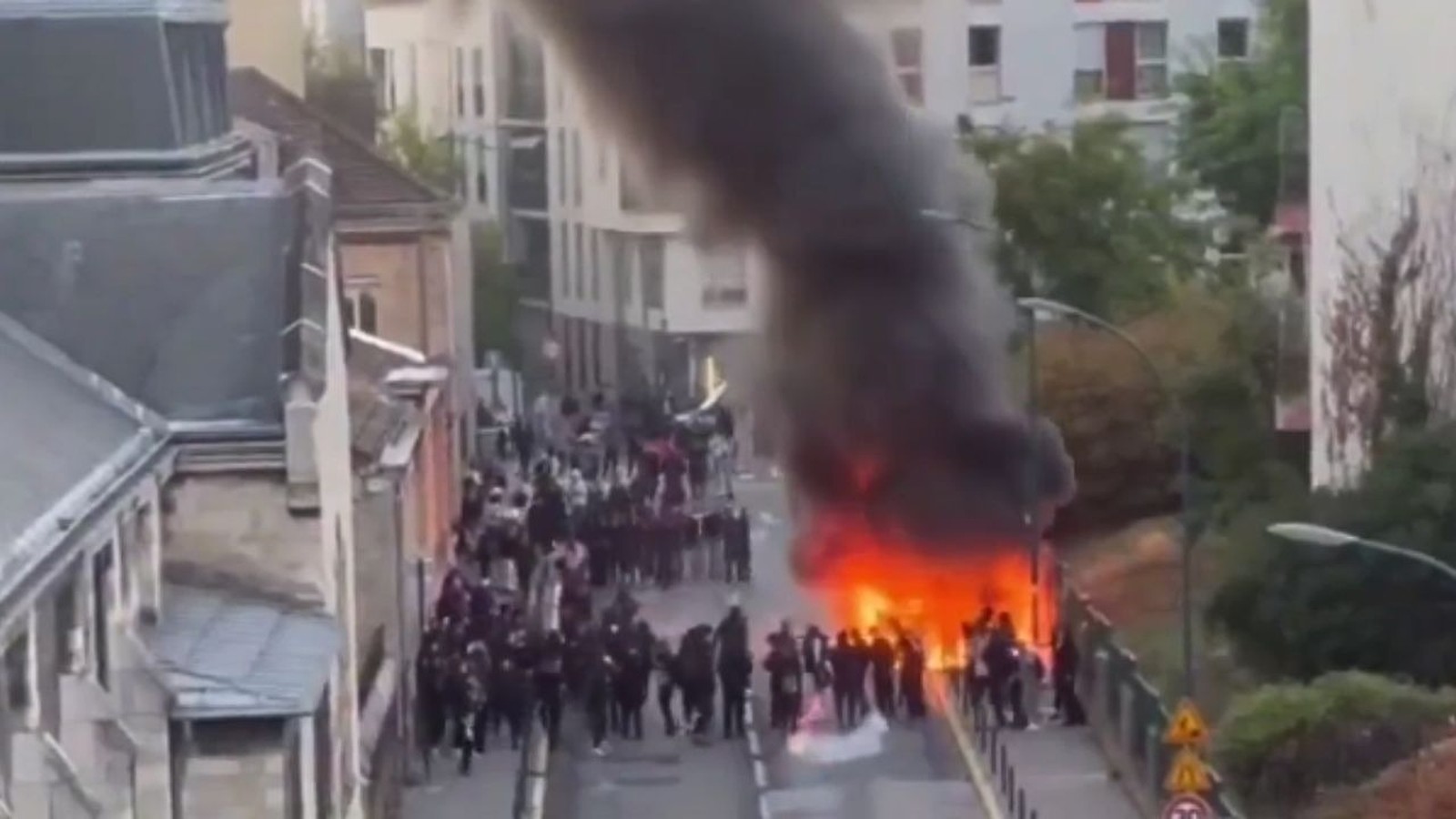 Vista aérea de uma rua com protesto estudantil, grande incêndio e fumaça escura subindo.