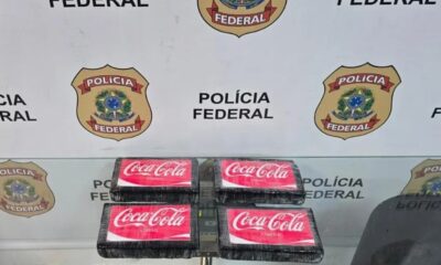 Quatro pacotes pretos com adesivos Coca-Cola classic sobre mesa, fundo Polícia Federal
