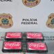 Quatro pacotes pretos com adesivos Coca-Cola classic sobre mesa, fundo Polícia Federal