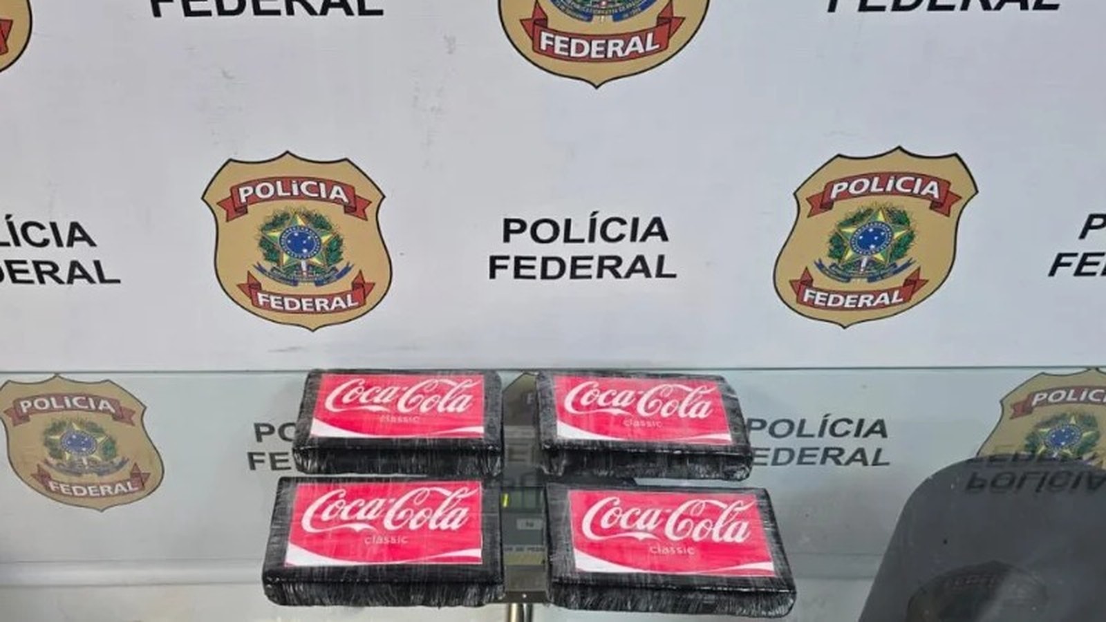 Quatro pacotes pretos com adesivos Coca-Cola classic sobre mesa, fundo Polícia Federal