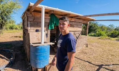 Jovem de camisa azul com 'WND NIG' posa ao lado de casa rústica de madeira e barril azul