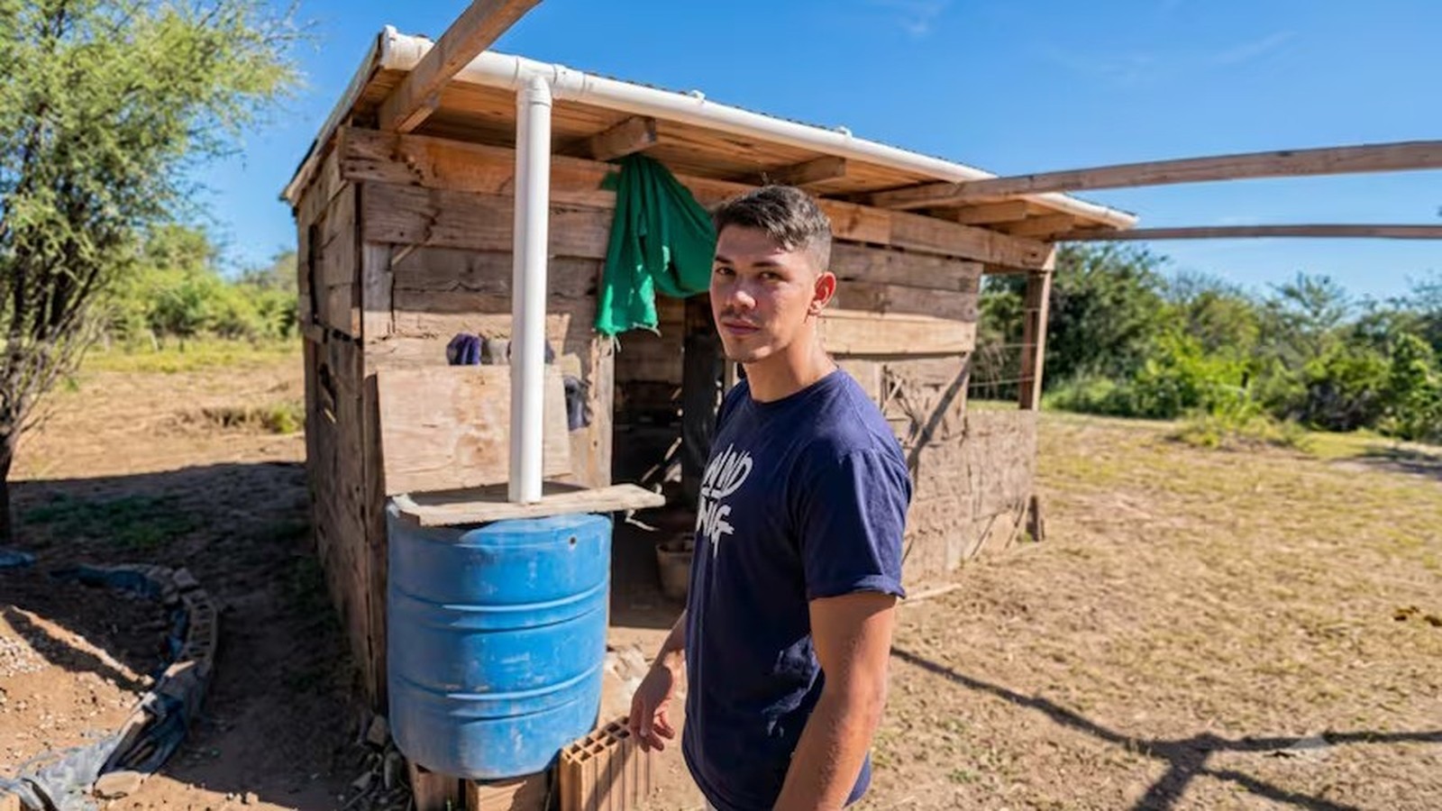 Jovem de camisa azul com 'WND NIG' posa ao lado de casa rústica de madeira e barril azul